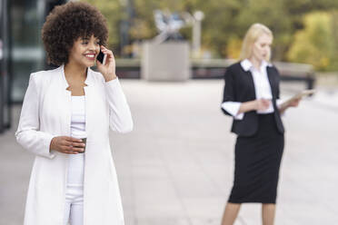 Businesswoman talking on mobile phone while standing with colleague in background on footpath - GGGF00127