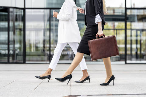 Businesswoman with briefcase using mobile phone while walking by colleague on footpath stock photo