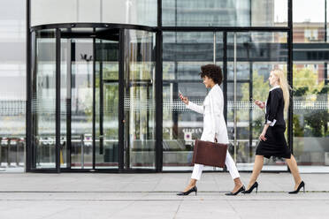 Businesswomen using mobile phone while walking with colleague against building - GGGF00143