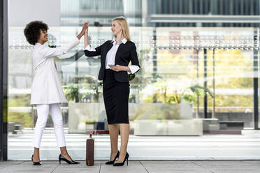 Businesswoman with mobile phone giving high five to colleague while standing against glass wall - GGGF00145