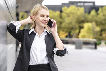 Lächelnde Geschäftsfrau, die wegschaut, während sie mit einem Handy in der Stadt telefoniert - GGGF00147