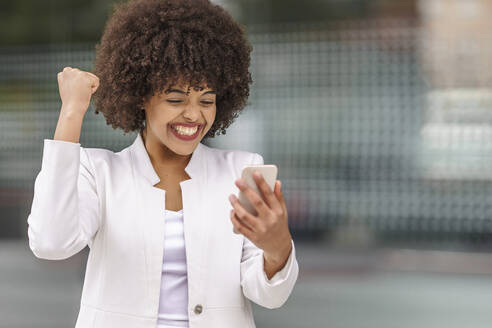 Cheerful businesswoman showing winning gesture while using mobile phone outdoors - GGGF00155