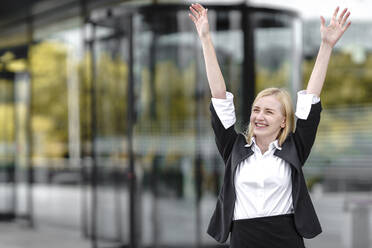 Smiling young woman with hand raised standing against building - GGGF00160