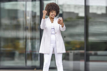 Junge Geschäftsfrau, die eine gewinnende Geste zeigt, während sie ein Mobiltelefon benutzt und im Freien steht - GGGF00161