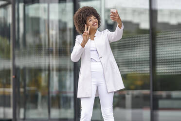 Lächelnde Geschäftsfrau, die eine Friedensgeste zeigt, während sie ein Selfie mit ihrem Mobiltelefon im Freien macht - GGGF00162