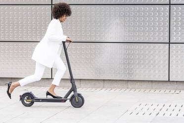 Smiling businesswoman riding electric push scooter against silver wall - GGGF00173