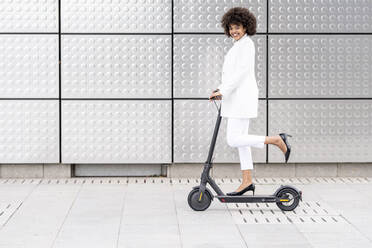 Businesswoman riding electric push scooter against silver wall - GGGF00174