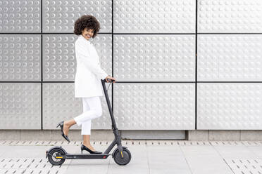 Businesswoman smiling while riding electric push scooter against silver wall - GGGF00175