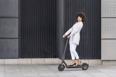 Excited businesswoman riding electric push scooter against wall - GGGF00176