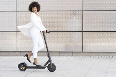 Smiling businesswoman looking away while riding push scooter against wall - GGGF00179