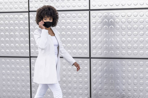 Businesswoman wearing face mask using mobile phone while walking against silver wall stock photo