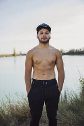 Shirtless young man with hands in pockets standing against lake and clear sky - ACPF00875