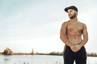 Young man wearing cap looking away while standing against clear sky and lake - ACPF00876