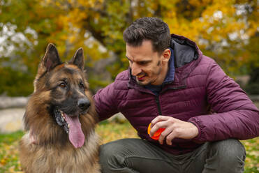 Lächelnder Mann mit Spielzeug, der seinen Hund anschaut, während er sich im Park hinhockt - OCMF01844
