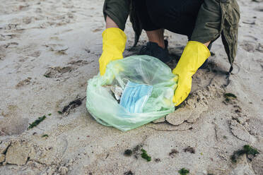 Woman collecting disposable face mask in garbage bag while cleaning beach - OYF00255