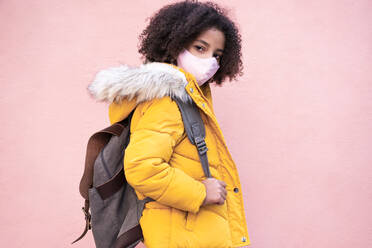 Girl with face mask carrying backpack while standing against pink wall - EBBF01410