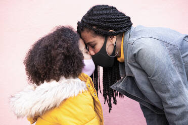 Mutter und Tochter mit Gesichtsmaske stehen von Angesicht zu Angesicht vor einer rosa Wand - EBBF01413