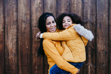 Mother carrying daughter while standing against wooden wall - EBBF01418