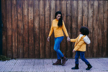 Mother holding daughter hand while walking against wooden wall - EBBF01420