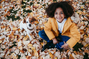 Smiling girl sitting with dog on fallen leaf at park - EBBF01433