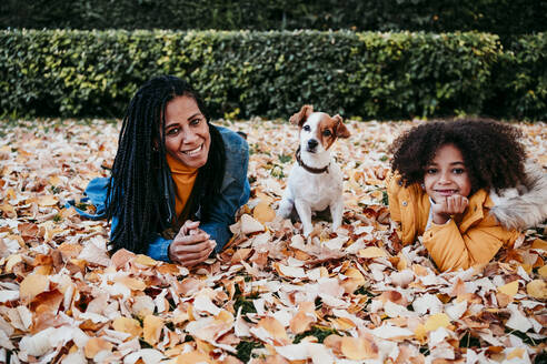 Smiling woman and daughter lying on dry leaf by dog sitting at park - EBBF01434