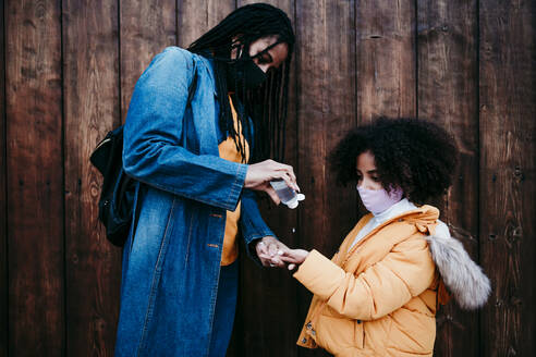 Mother giving alcohol hand sanitizer to daughter while standing against wooden wall - EBBF01440