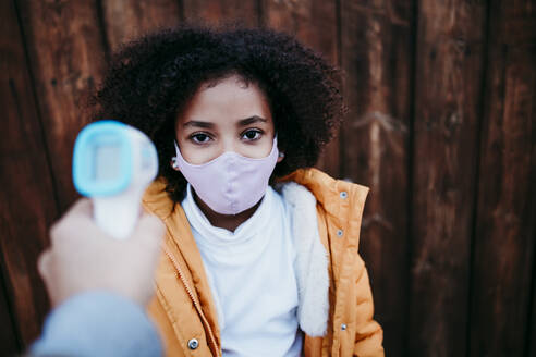 Mother using infrared thermometer while checking daughter temperature against wooden wall - EBBF01443