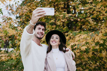 Smiling man taking selfie with female partner while standing against tree during autumn - EBBF01463