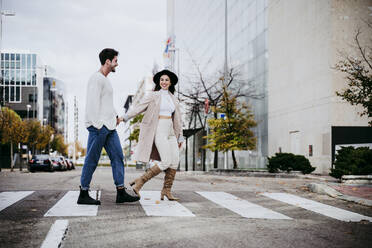 Smiling woman holding hands with male partner while crossing road in city - EBBF01466