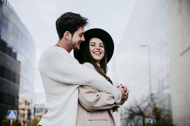 Smiling man embracing while looking at female partner against sky in city - EBBF01473