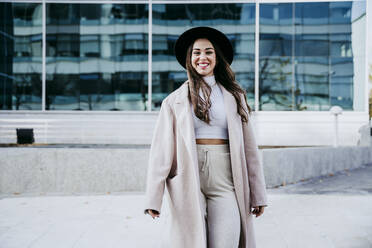 Beautiful smiling woman wearing hat while standing against building in city - EBBF01483