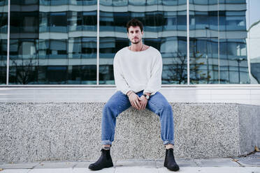 Confident man sitting on retaining wall against building in city - EBBF01486