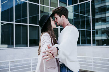 Smiling woman holding hands with male partner while kissing against building in city - EBBF01491