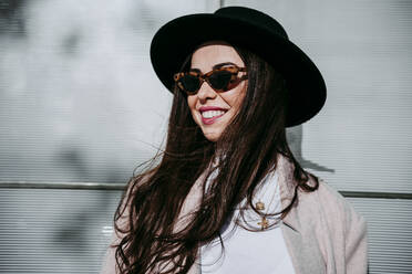 Beautiful smiling woman wearing hat while standing against wall - EBBF01499