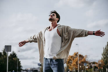 Confident man with arms outstretched against sky during sunny day - EBBF01504