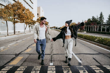 Beautiful smiling woman holding hands with male partner while walking on road in city - EBBF01507