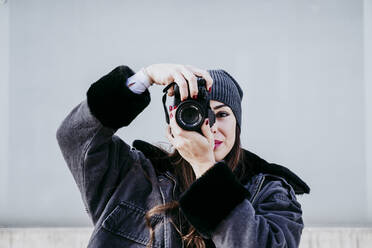 Confident woman taking photograph through camera while standing against wall - EBBF01523