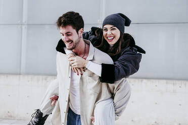 Smiling man piggybacking female partner while standing against wall - EBBF01529