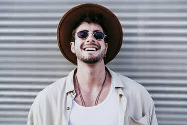 Happy man in hat and sunglasses standing against wall - EBBF01538