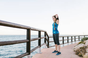 Seitenansicht einer fitten Frau in Sportkleidung, die auf einer Promenade steht und die Arme beim Training am Meer streckt - ADSF17284