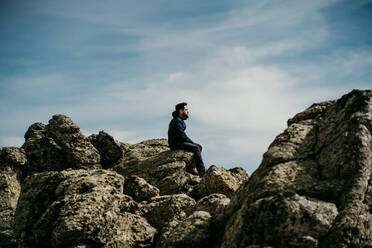 Seitenansicht eines Mannes, der mit geschlossenen Augen auf einem Stein sitzt und die Bergkette Puerto de la Morcuera an einem bewölkten Tag in Spanien bewundert - ADSF17301