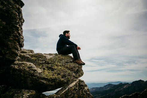 Seitenansicht eines Mannes, der am Rande eines Steins sitzt und die Bergkette Puerto de la Morcuera an einem bewölkten Tag in Spanien bewundert - ADSF17302