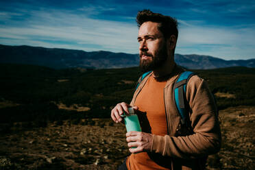 Side view of bearded man with bottle of water standing on stone against overcast sky during trip in Puerto de la Morcuera mountains in Spain - ADSF17305