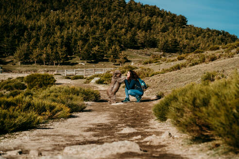 Seitenansicht von glücklichen weiblichen lächelnd hockte mit flauschigen Labradoodle beim Spaziergang entlang des Weges an einem sonnigen Tag in Puerto de la Morcuera Berge in Spanien - ADSF17307