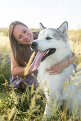 Delighted female in summer dress and straw hat relaxing on meadow with Alaskan Malamute and looking at camera - ADSF17318