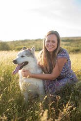 Delighted female in summer dress and straw hat relaxing on meadow with Alaskan Malamute and looking at camera - ADSF17319