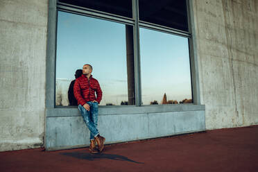 Selbstbewusster junger unrasierter Mann in roter warmer Jacke und Jeans, der wegschaut, während er steht und sich gegen ein großes Fenster lehnt - ADSF17351