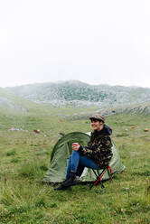 Full length content female traveler in jeans and warm khaki jacket sitting on folding chair near camping tent with cup of hot drink during camping in highlands - ADSF17353