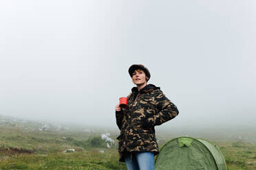 Von unten zufriedene weibliche Reisende in Jeans und warmer khakifarbener Jacke, die mit einer Tasse Heißgetränk beim Camping im nebligen Hochland in der Nähe des Zeltes steht - ADSF17354