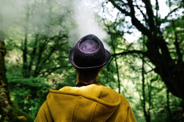 Back view of unrecognizable female traveler with hooded yellow coat exhaling smoke and looking over shoulder on blurred background of green forest - ADSF17360